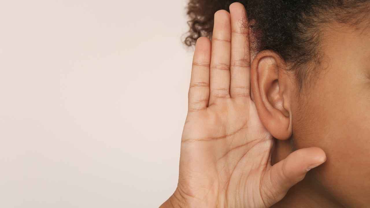 Close no ouvido de uma pessoa com a mão posicionada atrás da orelha em gesto de escuta atenta.