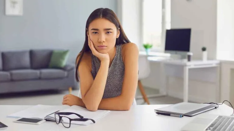 Mulher com expressão de frustração sentada em escritório, com materiais de trabalho à sua frente, sugerindo bloqueio criativo causado por ambiente barulhento.
