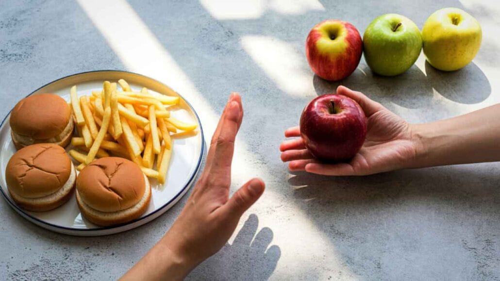 Mãos segurando uma maçã vermelha ao lado de prato com hambúrgueres e batatas fritas, representando escolha alimentar.