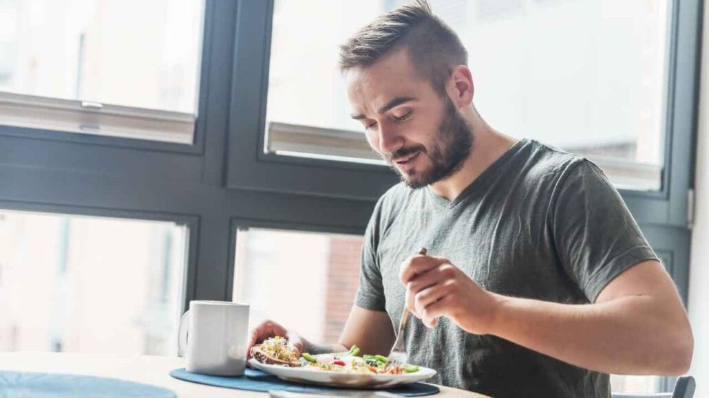 Homem sentado à mesa próximo a uma janela antirruído comendo prato com salada e acompanhamentos, com uma caneca ao lado.