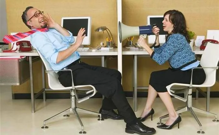 Mulher usando um megafone para falar com colega de trabalho em escritório, enquanto o homem se afasta incomodado com o som alto, representando o impacto negativo do barulho no ambiente corporativo.