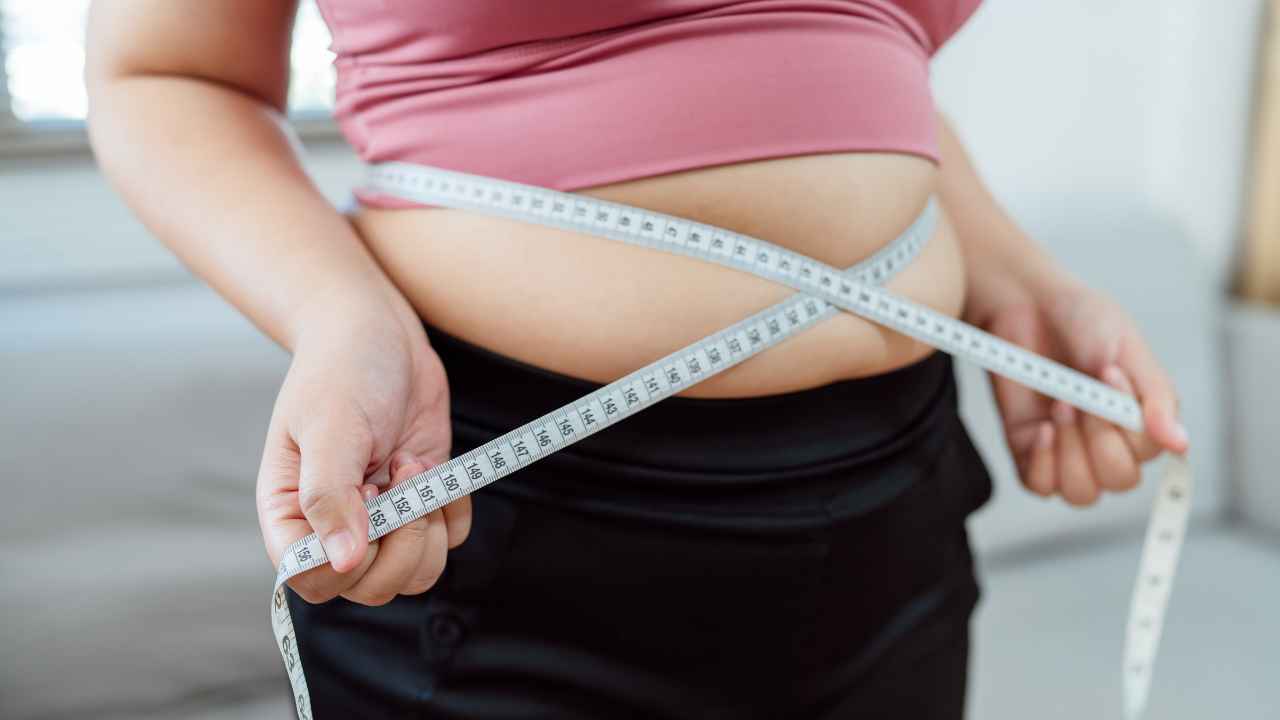 Mulher usando fita métrica para medir a circunferência da cintura, com foco na região abdominal e nas mãos segurando a fita.