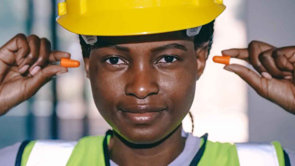 Mulher usando capacete de segurança amarelo e colete refletivo segura um protetor auricular de espuma laranja em cada mão, pronta para colocá-los nos ouvidos.