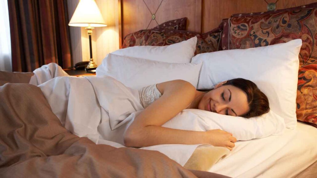 Mulher dormindo tranquilamente em uma cama de hotel, com expressão serena e ambiente acolhedor ao redor.