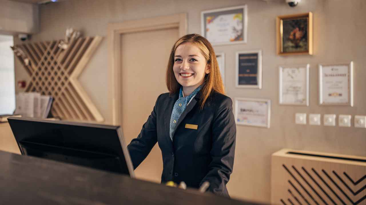 Recepcionista sorridente em balcão de hotel, com certificados desfocados na parede ao fundo, representando o selo Quiet Room.