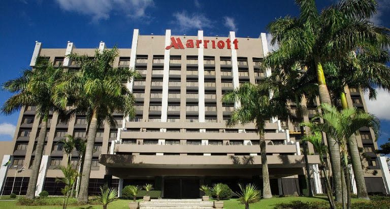 Fachada do hotel Marriott em meio a um jardim com palmeiras, sob um céu azul com poucas nuvens.