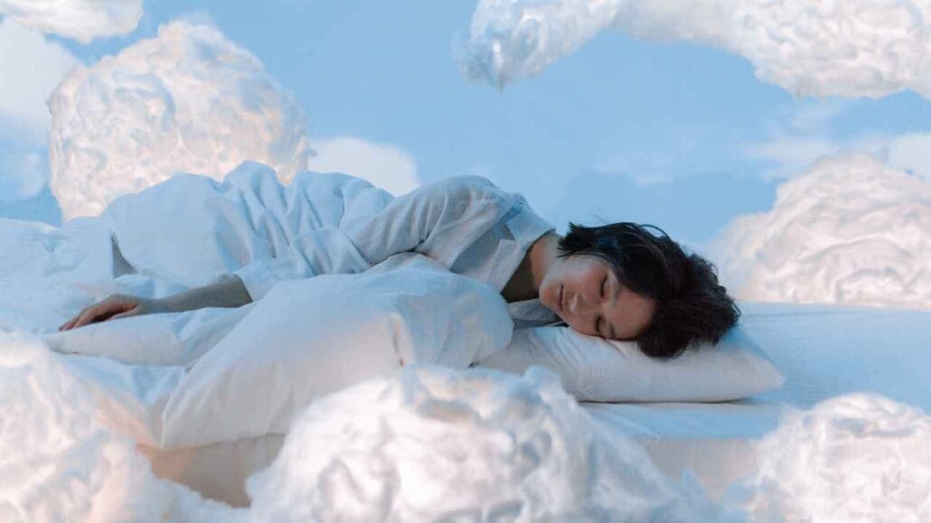 Mulher dormindo tranquilamente em uma cama branca cercada por nuvens, representando o estágio de sono REM