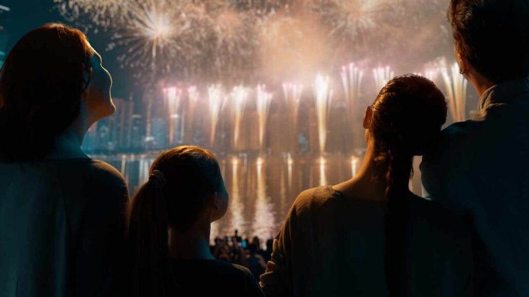 Família reunida à beira da água observando á distância um grande espetáculo de fogos de artifício no céu noturno.