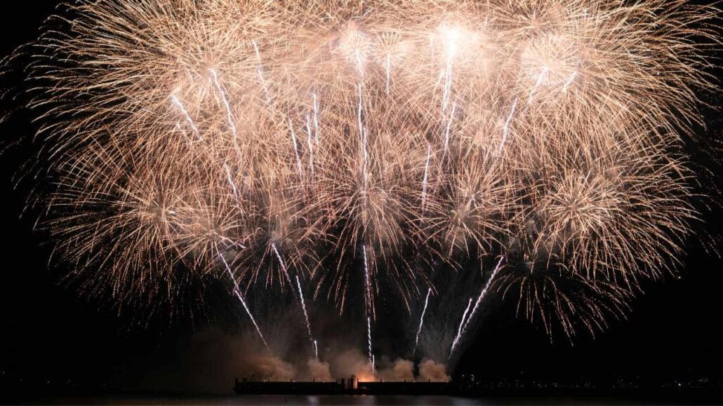 Fogos de artifício estourando intensamente no céu, formando padrões amplos e luminosos durante uma celebração.
