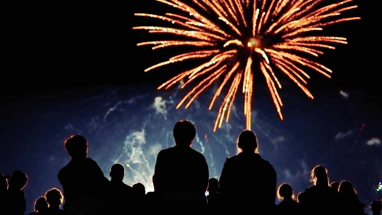 Grupo de pessoas observa fogos de artifício iluminando o céu noturno, com explosões coloridas ao fundo.