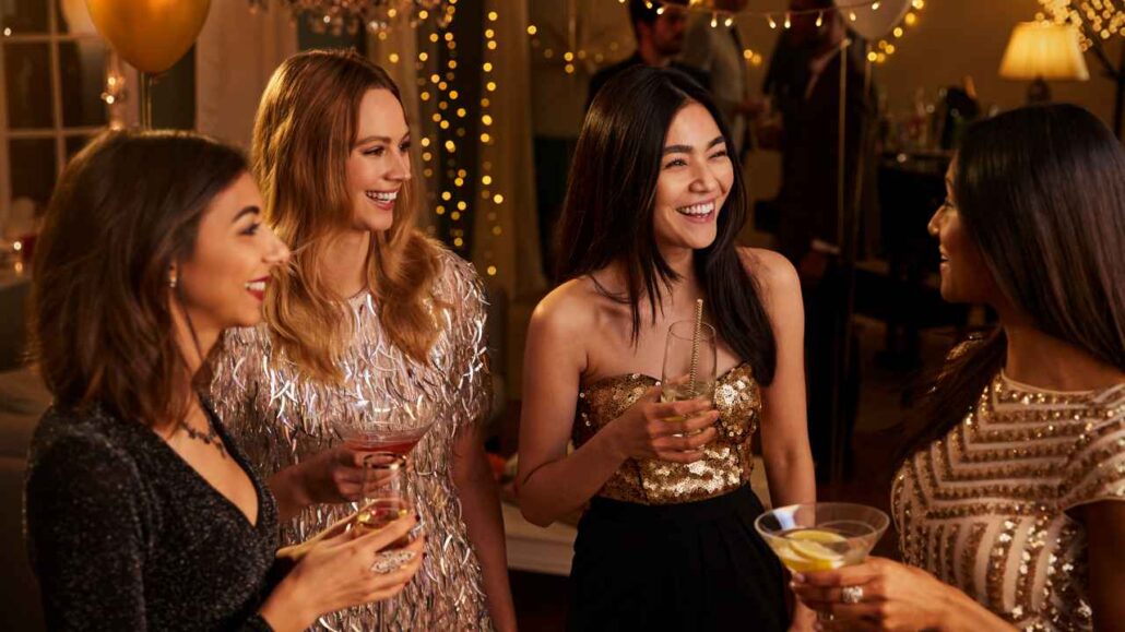 Grupo de quatro mulheres sorrindo e conversando em um ambiente festivo decorado com luzes, segurando drinks e aproveitando um momento descontraído em um local calmo da festa.