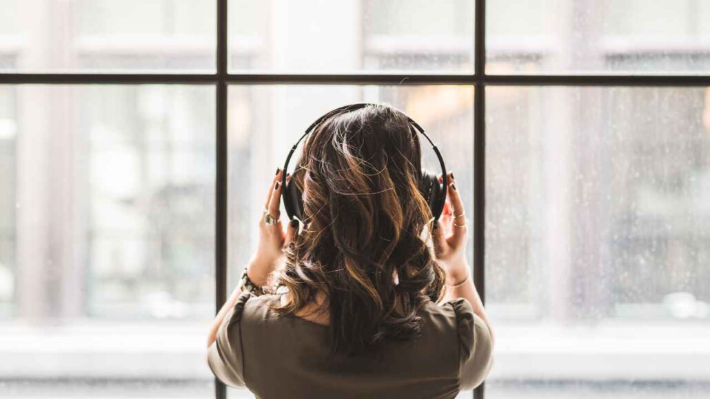 Mulher vista de costas, usando fones de ouvido enquanto olha pela janela, em um momento de concentração e relaxamento ao escutar ruído branco para abafar sons externos.