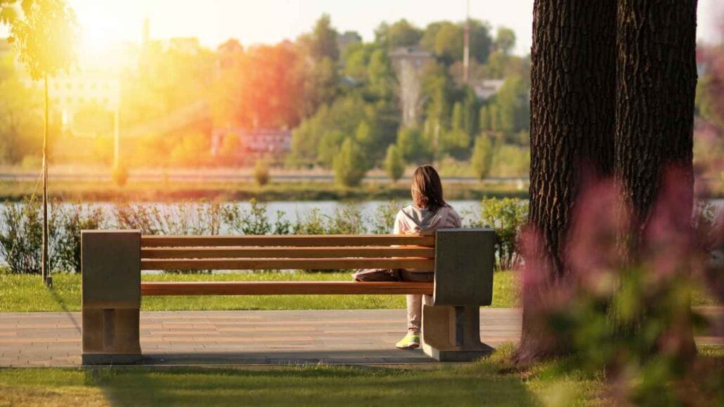 Mulher sentada sozinha em um banco de parque com vista para um lago e vegetação abundante ao entardecer