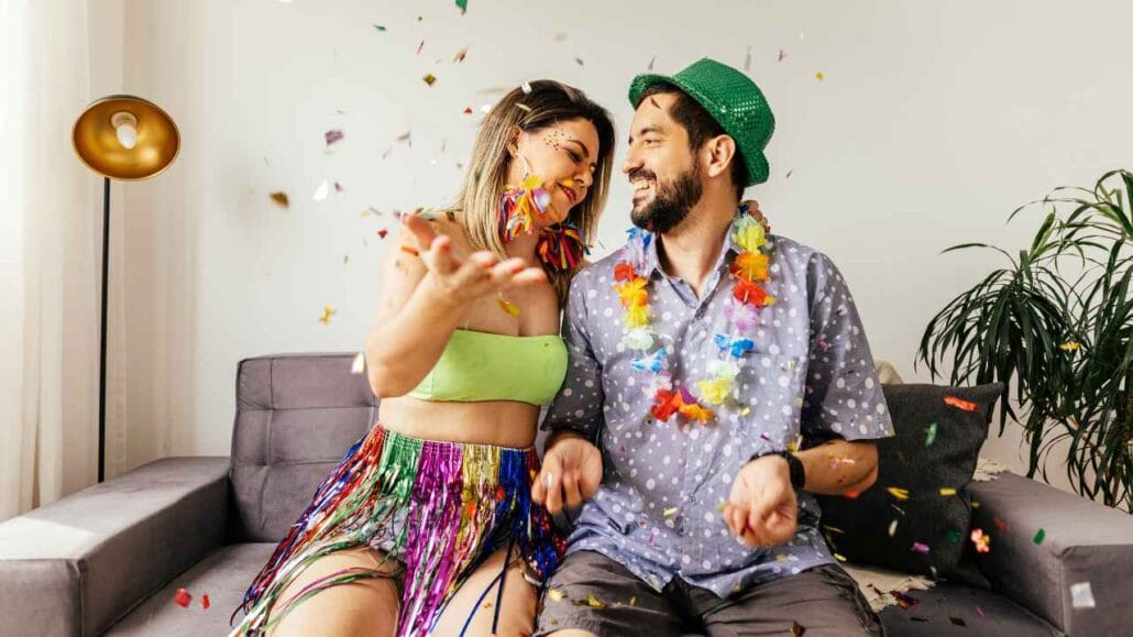 Casal usando adereços carnavalescos comemora em casa, jogando confetes e celebrando o Carnaval em um ambiente silencioso