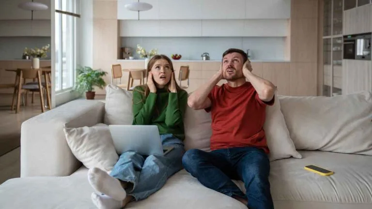 Casal sentado no sofá da sala com expressão de incômodo, cobrindo os ouvidos com as mãos enquanto olha para o teto. A imagem transmite desconforto causado por ruídos residenciais vindos do andar de cima.
