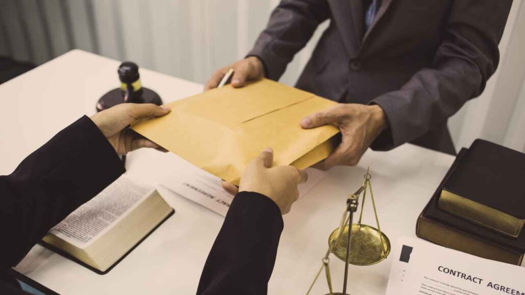 Duas pessoas trocando um envelope pardo sobre uma mesa com livros jurídicos, contrato e balança da justiça.