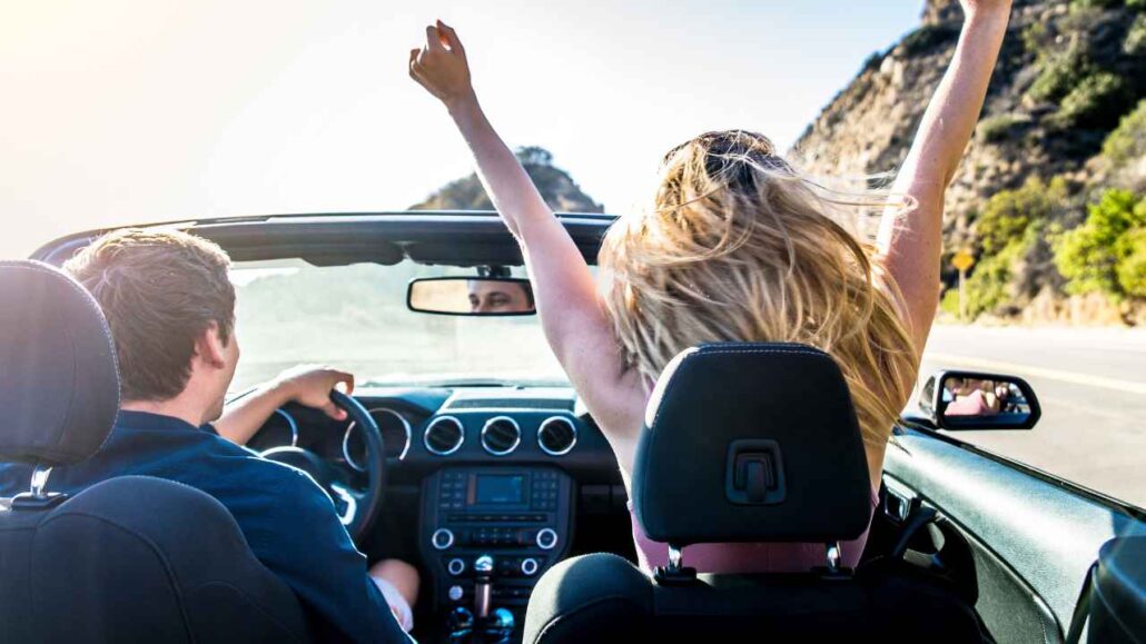 Duas pessoas em um carro conversível em movimento por uma estrada ensolarada, transmitindo sensação de liberdade e diversão durante a viagem.