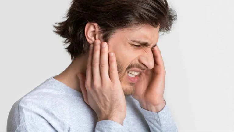 Homem jovem com expressão de dor segurando as duas laterais da cabeça, pressionando as orelhas devido a um desconforto auditivo.