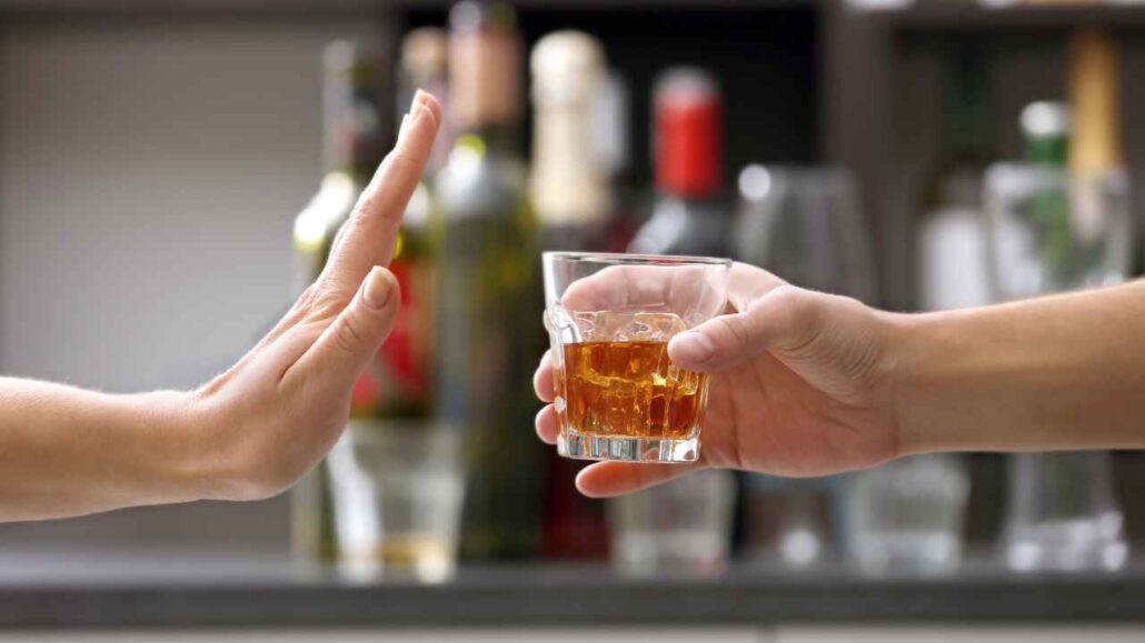 Mão de uma pessoa fazendo sinal de recusa enquanto outra oferece um copo com bebida alcoólica em um bar.