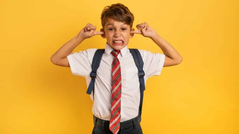 Criança com uniforme escolar branco, gravata listrada vermelha e mochila azul, tampa os ouvidos com os dedos e faz expressão de incômodo em frente a um fundo amarelo.
