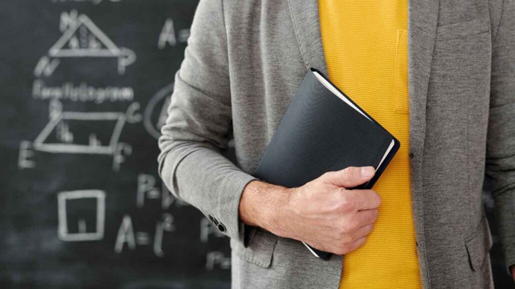 Homem usando blazer cinza e camiseta amarela segura um caderno preto em frente a um quadro negro com fórmulas e figuras geométricas desenhadas, representando o ambiente escolar.