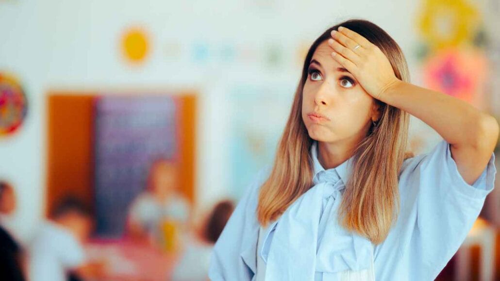 Professora jovem, com expressão de cansaço e mão na testa, parece sobrecarregada em sala de aula, enquanto alunos desfocados aparecem ao fundo.