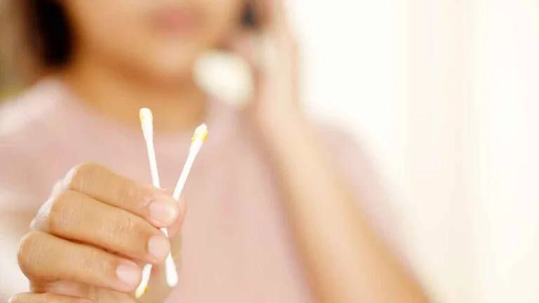 Pessoa segura dois cotonetes com cera de ouvido visível nas pontas, ilustrando o uso do acessório para limpeza auricular.