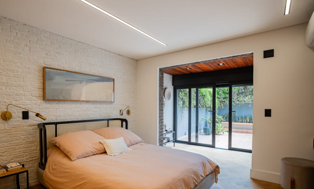Quarto moderno com cama de casal, roupa de cama em tom bege, parede de tijolos brancos aparentes e portas antirruido ao fundo que dão acesso a uma área externa com plantas.
