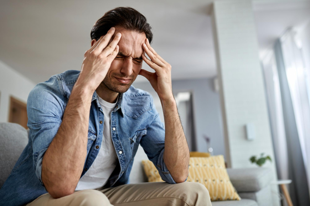 Homem sentado em casa com expressão de dor e as mãos na cabeça, aparentando incômodo intenso devido aos ruídos do dia a dia, como trânsito, obras ou vizinhança barulhenta.
