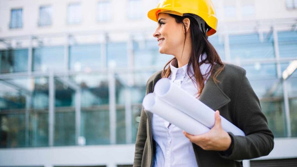 Engenheira usando capacete amarelo segura rolos de plantas na área externa de um edifício moderno