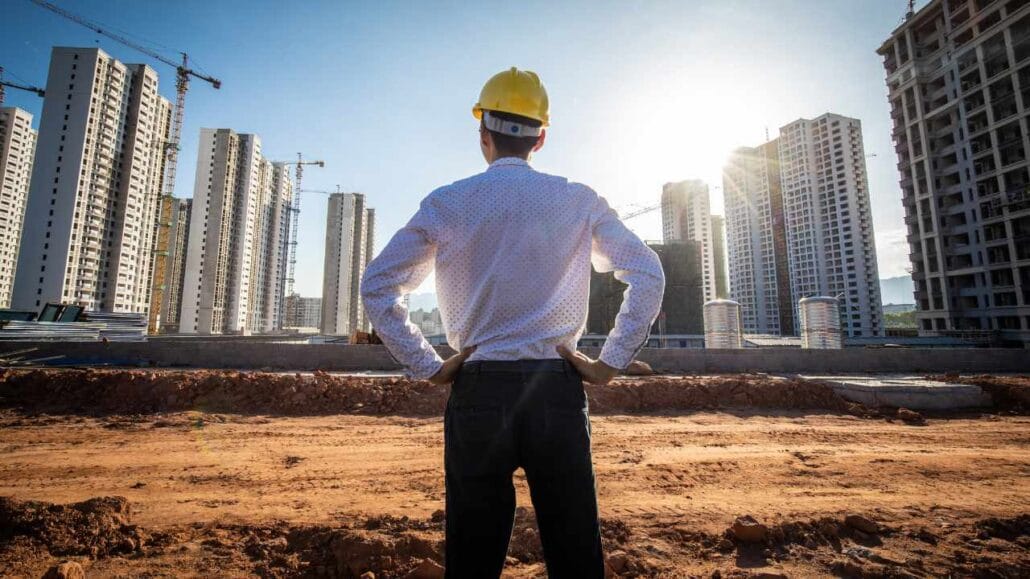 Homem com capacete amarelo observa um grande canteiro de obras ao ar livre, com diversos prédios altos em construção ao fundo
