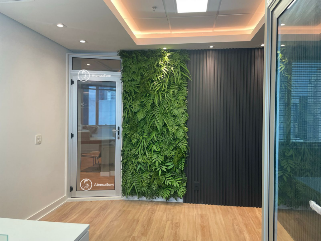 Porta de vidro com isolamento acústico, com logotipo da Atenua Som, ao lado de uma parede verde vertical decorativa e painel preto ripado em um ambiente moderno com piso de madeira.