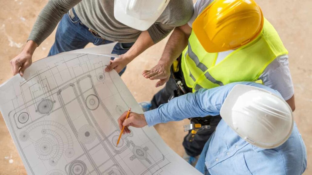 Grupo de profissionais da construção civil com capacetes revisa e aponta detalhes em uma planta arquitetônica aberta sobre a mesa
