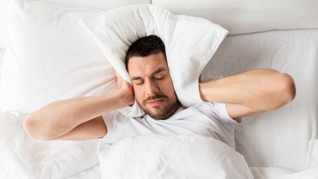 Homem incomodado com barulho e não conseguindo dormir. Ele tapa as orelhas com o travesseiro.