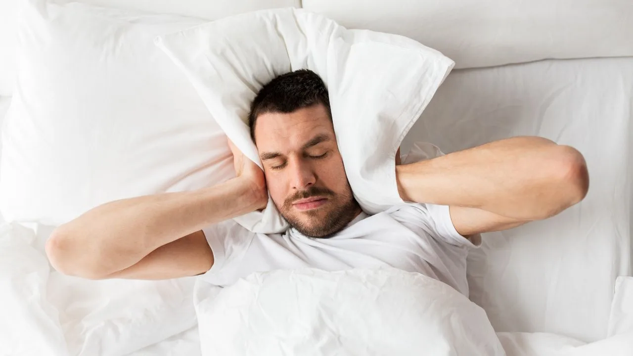 Homem incomodado com barulho e não conseguindo dormir. Ele tapa as orelhas com o travesseiro.