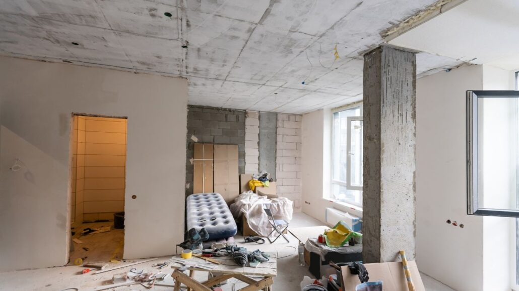 Interior de um apartamento em reforma, com paredes parcialmente rebocadas, piso coberto por ferramentas e materiais, colchão inflável e móveis temporários, próximo a janelas abertas.