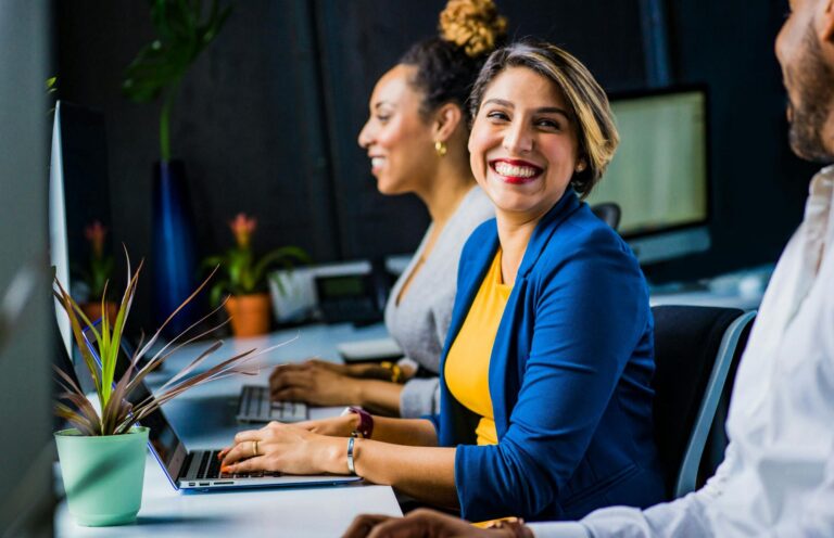 Three diverse professionals working and smiling at office desks, fostering teamwork and collaboration.
