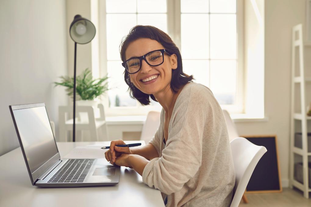 Mulher sorridente, usando óculos, sentada à frente de um notebook em uma mesa branca, segurando uma caneta. Ao fundo, ambiente iluminado por luz natural, com planta decorativa e janela grande.