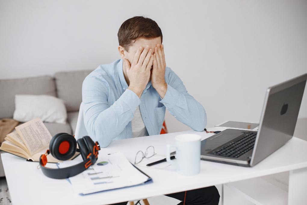 Homem sentado à mesa de trabalho em home office, vestindo camisa azul clara, cobrindo o rosto com as mãos em sinal de cansaço ou frustração. À frente, há um notebook, caneca azul clara, óculos, fones de ouvido, gráficos impressos e um livro aberto.
