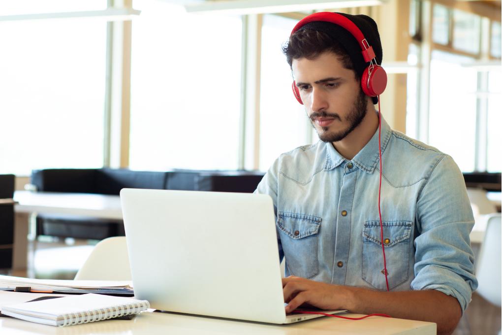 Homem jovem usando camisa jeans e fones de ouvido vermelhos, concentrado no notebook em ambiente claro e moderno, com caderno e caneta sobre a mesa.