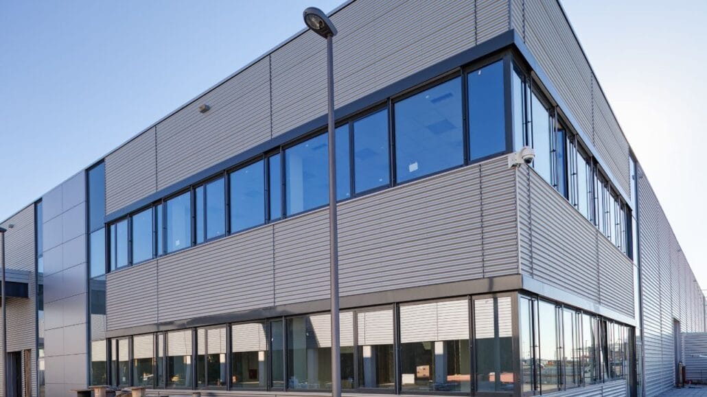 Fachada de um prédio industrial moderno com revestimento metálico e amplas janelas de vidro refletivo, sob céu limpo e iluminação natural.