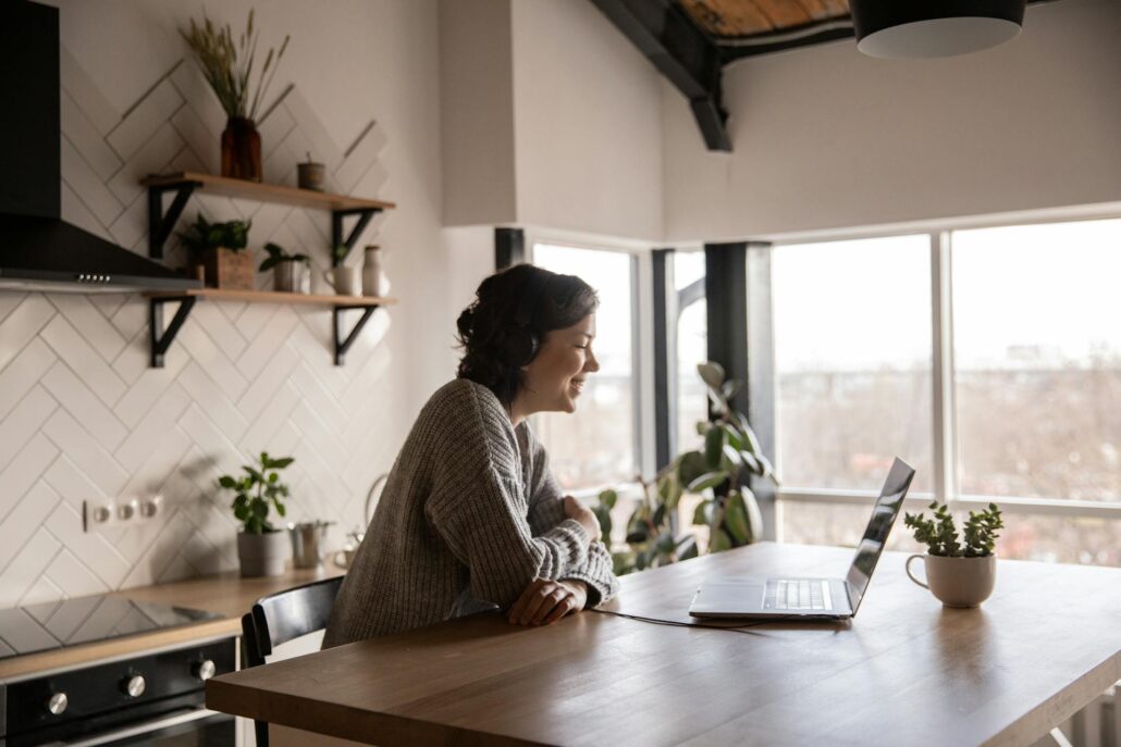Mulher sentada em uma cozinha iluminada, usando fones de ouvido e sorrindo para a tela de um notebook sobre a mesa, sugerindo trabalho em home office. Ao fundo, prateleiras com plantas e utensílios, e uma janela ampla que deixa entrar luz natural.