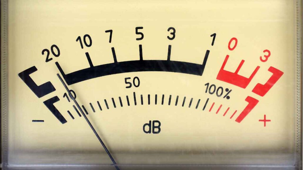 Medidor analógico de decibel com ponteiro indicando intensidade sonora próxima de 10 dB, com escala graduada em preto e vermelho que representa diferentes níveis de ruído.