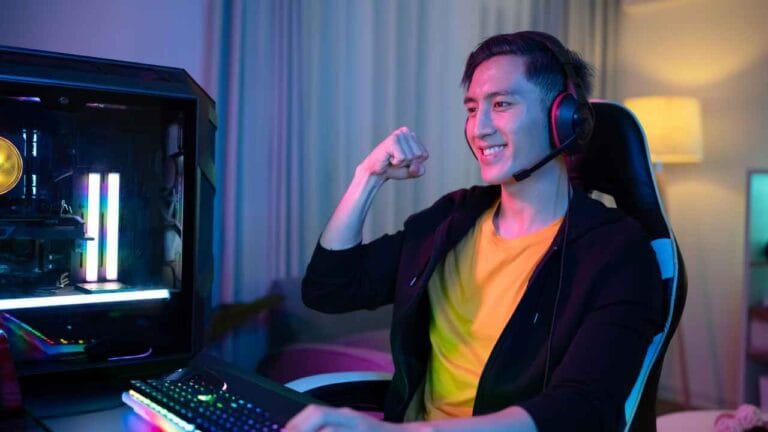 Jovem gamer sentado em cadeira ergonômica comemora com expressão de vitória enquanto joga no computador, usando headset com microfone e rodeado por iluminação LED colorida em ambiente doméstico.