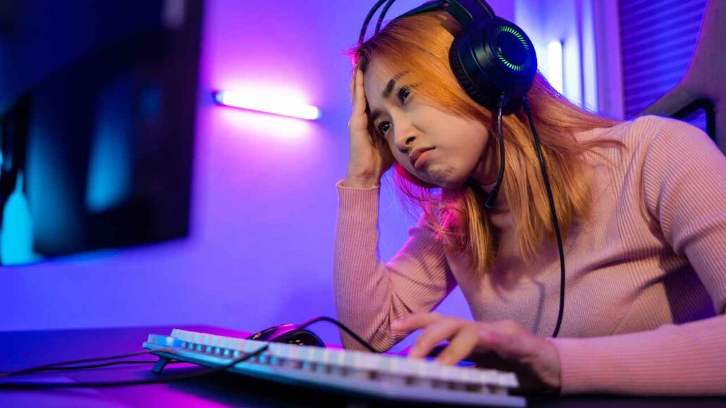 Jovem gamer com expressão de frustração segura a cabeça enquanto joga no computador, usando fone que não consegue bloquear o barulho do ambiente, dificultando sua concentração.