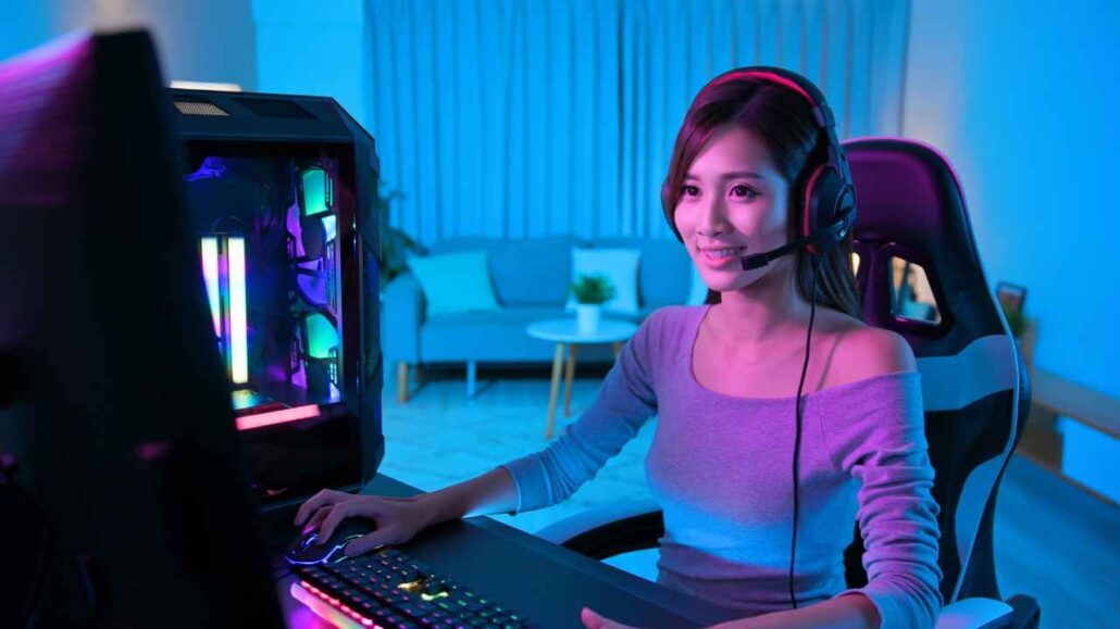 Mulher gamer com headset e microfone joga em computador com gabinete iluminado por luzes LED coloridas, sentada em cadeira ergonômica em ambiente moderno com iluminação azul.