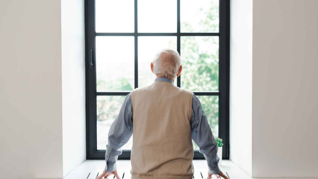 Homem idoso de costas, contemplando a paisagem pela janela. A estrutura da janela antirruído proporciona um ambiente tranquilo e silencioso dentro de casa.