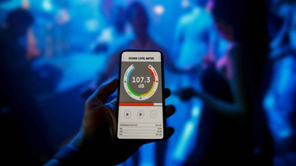 Pessoa segura um smartphone exibindo um aplicativo de medição de ruído com leitura de 107,3 decibel, em um ambiente escuro e iluminado por luzes azuis, sugerindo uma festa ou balada com som elevado.