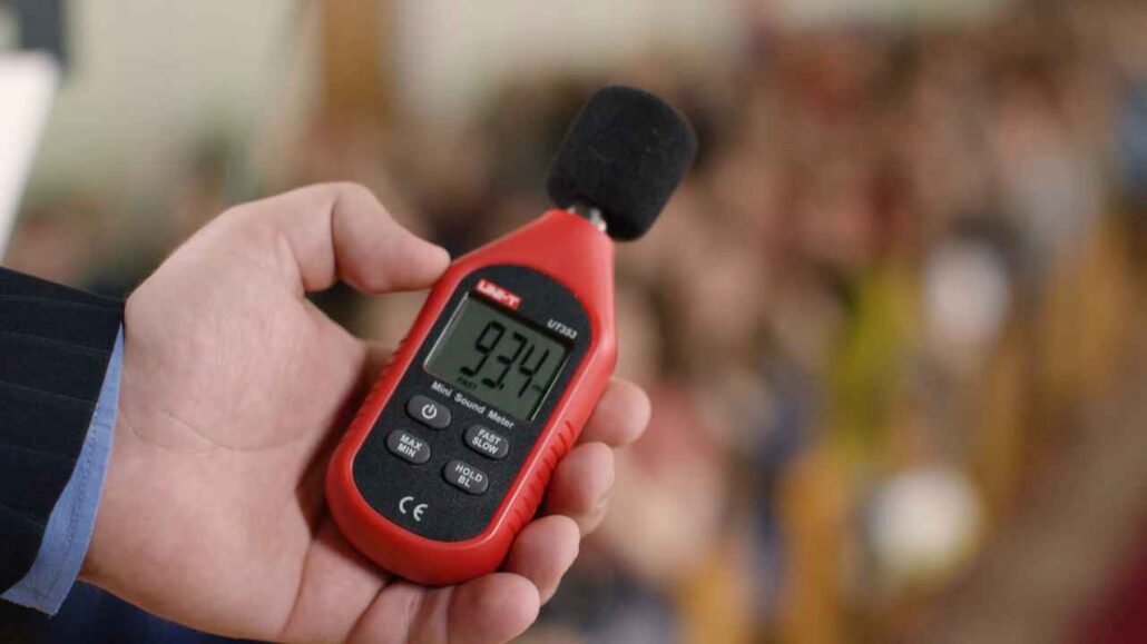 Pessoa segura um decibelímetro digital vermelho com visor exibindo a medição de 93,4 decibel, em um ambiente com fundo desfocado, indicando a análise do nível de ruído no local.