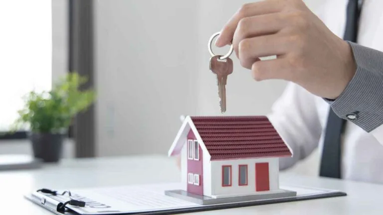 Mão de uma pessoa segurando uma chave sobre uma maquete de casa com telhado vermelho, posicionada sobre um contrato de compra ou aluguel em uma mesa branca.